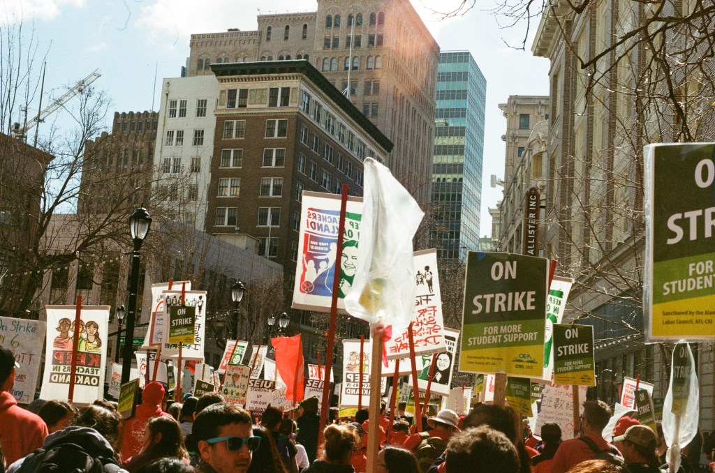 Photo of a crowd on Telegraph Avenue in downtown Oakland, California, during the OEA teachers' strike in 2019. Crowd members are holding green and yellow signs that read, "On strike for more student support," "Keep teachers in Oakland," and "Fight for the schools students deserve."